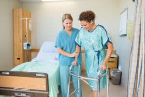 Nurse and woman in hospital gown using walker to move around after mommy makeover surgery.
