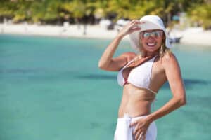 Smiling mature woman in a white bikini and sun hat standing by turquoise water.