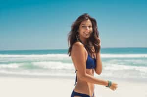 Smiling woman in a blue bikini walking along the beach, featured in a blog about breast augmentation and breast asymmetry.