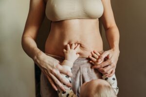 Mother holds baby’s hands against loose abdominal skin while considering mommy makeover